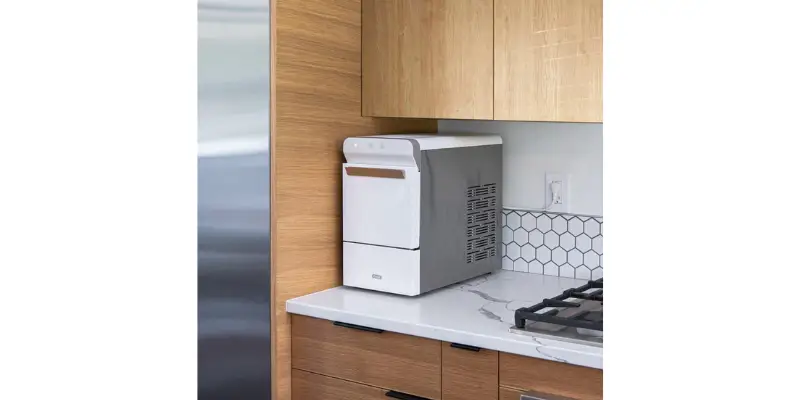 The Compact White And Silver Gevi Household V2.0 Nugget Ice Maker Sitting On A Modern Kitchen Countertop Next To A Gas Range. The Surrounding Cabinetry Is Light Wood