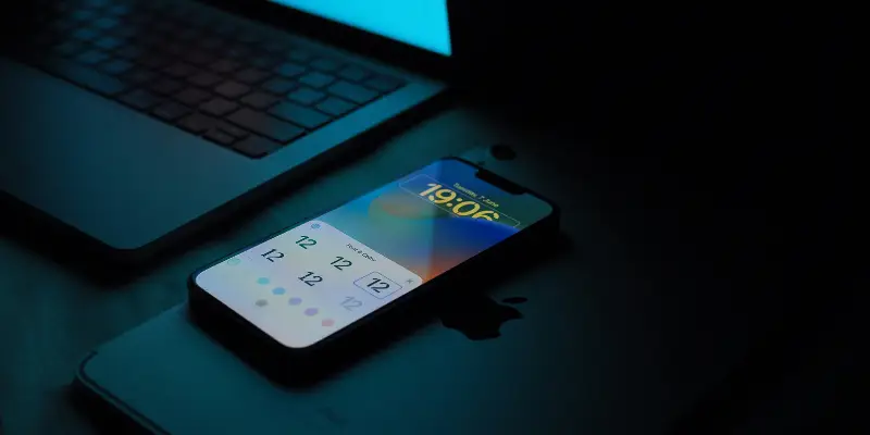 An Iphone Displaying A Customization Screen For The Lock Screen With Options For Clock Styles And Colors Resting On Another Apple Device Next To A Laptop In A Dimly Lit Setting With Bitbyteiq.com